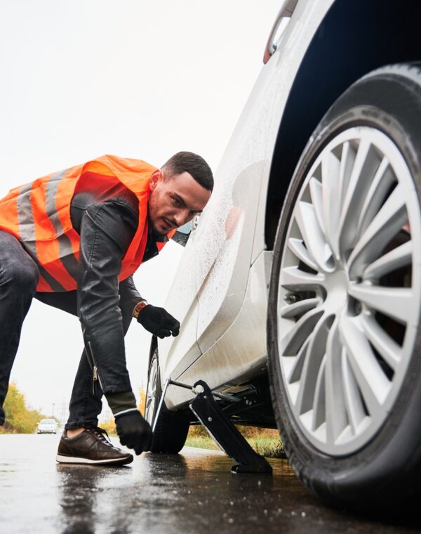 Roadside assistance worker raising vehicle with car jack before changing flat tire. Young man using special device for lifting car while repairing wheel on the road. Concept of emergency road service.