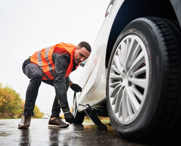 Roadside assistance worker raising vehicle with car jack before changing flat tire. Young man using special device for lifting car while repairing wheel on the road. Concept of emergency road service.