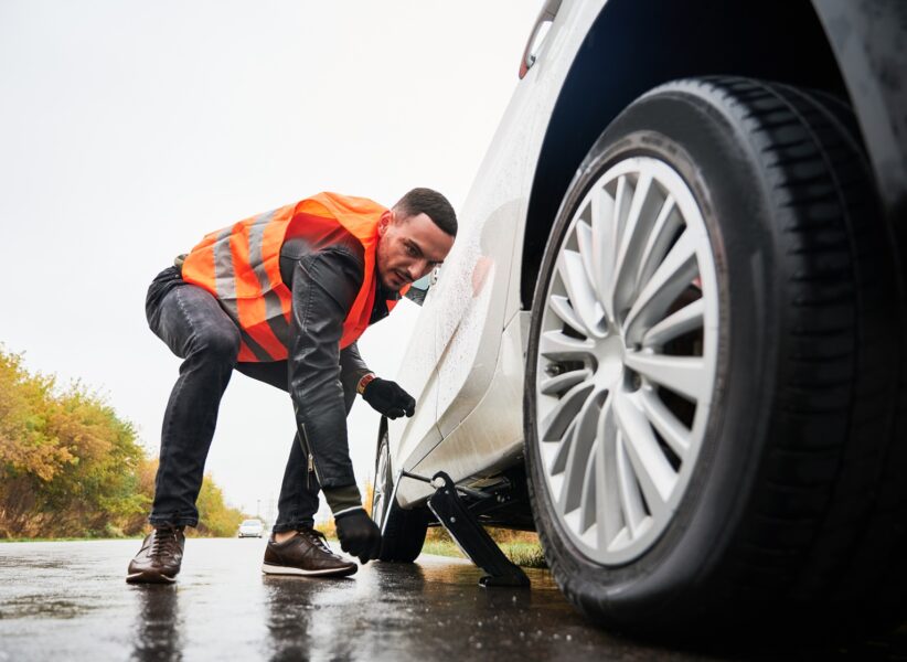 Roadside assistance worker raising vehicle with car jack before changing flat tire. Young man using special device for lifting car while repairing wheel on the road. Concept of emergency road service.