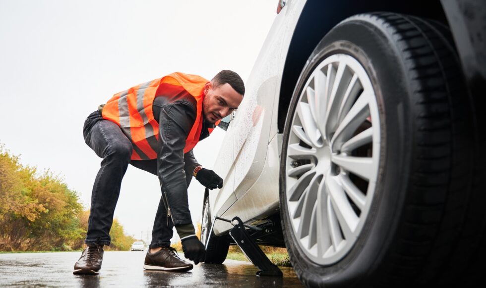 Roadside assistance worker raising vehicle with car jack before changing flat tire. Young man using special device for lifting car while repairing wheel on the road. Concept of emergency road service.